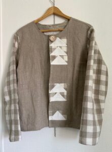 Handgjord jacka i naturfärgat tyg med rutiga ärmar och dekorativa vita triangulära applikationer på framsidan.