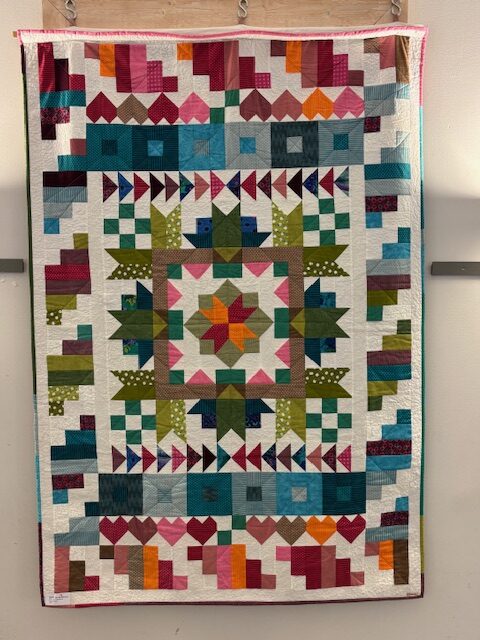 Handgjord quilt i färgstarkt geometriskt mönster med hjärtan, trianglar och block i grönt, blått, rosa och orange på vit botten.