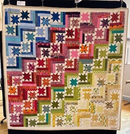 Handgjord quilt i många färger med stjärnblock och geometriska mönster i blått, rosa, grönt och gult, hängande på utställning.
