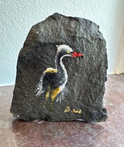 Målning av en häger med röd näbb, vit hals och gula inslag, målad direkt på en natursten.